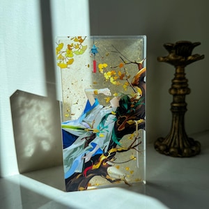 Peut inclure: Un bloc acrylique transparent avec une illustration de style anime d'une personne aux cheveux verts, v&ecirc;tue d'une tenue blanche et bleue, allong&eacute;e sur une branche d'arbre. L'&oelig;uvre comprend des feuilles jaunes et une lanterne suspendue. Un chandelier dor&eacute; est &agrave; droite.