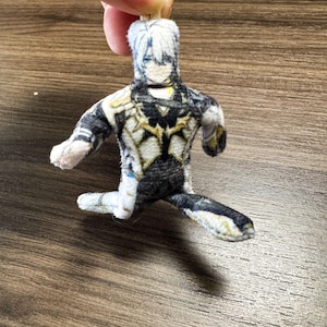 Op de afbeelding: Een kleine pluche pop in de vorm van een personage met wit haar en een gedetailleerde outfit in zwart, wit en goud. De pop zit met &eacute;&eacute;n been uitgestrekt en armen uitgestrekt. De pop wordt door een vinger vastgehouden.