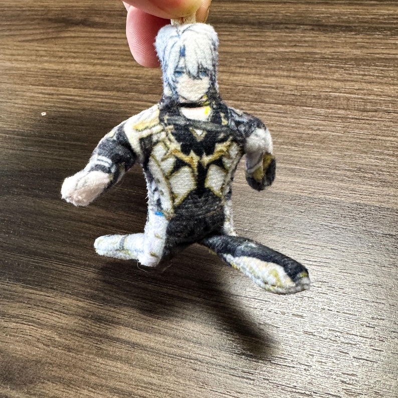 Op de afbeelding: Een kleine pluche pop in de vorm van een personage met wit haar en een gedetailleerde outfit in zwart, wit en goud. De pop zit met &eacute;&eacute;n been uitgestrekt en armen uitgestrekt. De pop wordt door een vinger vastgehouden.