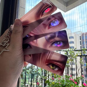 Puede incluir: Un conjunto de cuatro marcapáginas rectangulares con ilustraciones en primer plano de ojos de estilo anime con colores vibrantes. Los ojos tienen iris rojos, azules, morados y verdes. Un llavero plateado con un dije de estrella está adjunto.