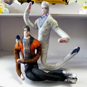 May include: Two handmade fabric dolls. One doll is in a white suit with a blue shirt, and the other is wearing an orange jacket, grey shirt, and black trousers. The dolls are on a white shelf.