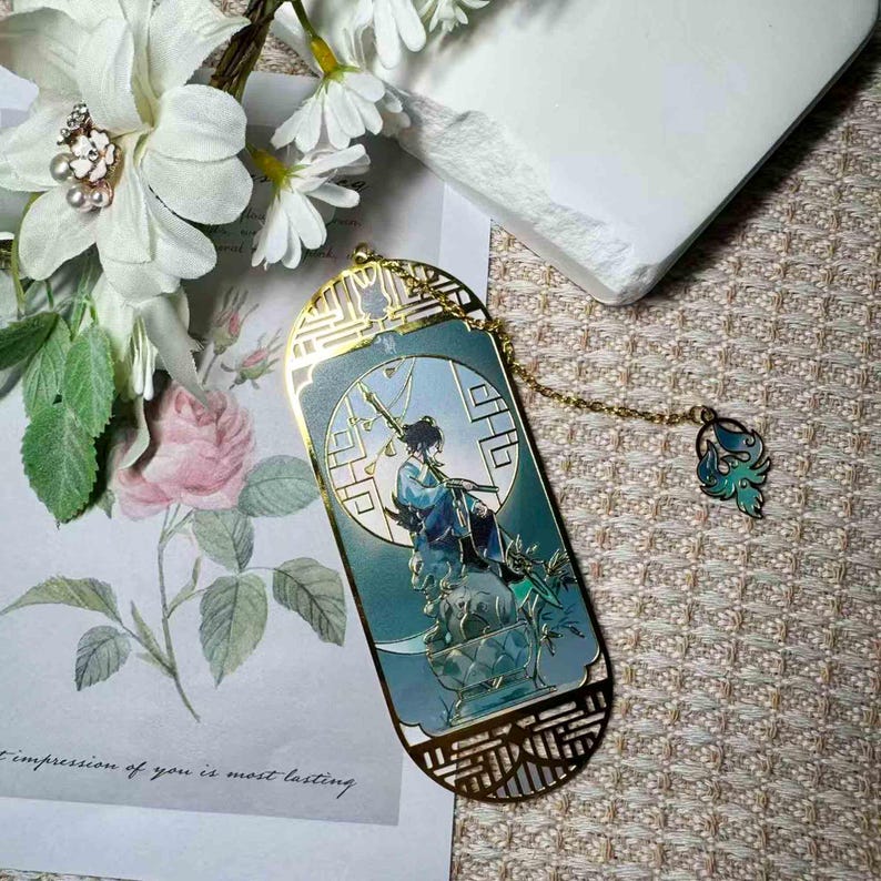May include: A gold-colored metal bookmark with a teal and blue illustration of a figure with a sword. The bookmark has a decorative chain and charm. The background is a soft, patterned surface with flowers.