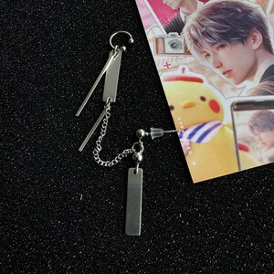 Op de afbeelding: Een paar zilverkleurige oorbellen. E&eacute;n oorbel heeft een gebogen bovenkant met twee rechthoekige staven en een ketting. De andere heeft een pin met een kleine bal en een rechthoekige staaf. Op een zwarte ondergrond.