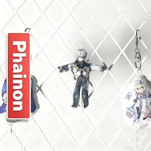 Op de afbeelding: Een verzameling sleutelhangers met anime-stijl personages. E&eacute;n sleutelhanger heeft het woord "Phainon" in het wit op een rode achtergrond. Een andere sleutelhanger is een kleine popachtige figuur met uitgestrekte armen. De derde sleutelhanger heeft een stripfiguur met een cadeau.