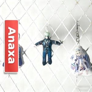 Op de afbeelding: Een rode rechthoekige tag met de witte tekst "Anaxa" wordt getoond naast twee sleutelhangers met personages. De sleutelhangers tonen personages in anime-stijl in blauwe en witte outfits. De achtergrond is een wit ruitvormig rooster.