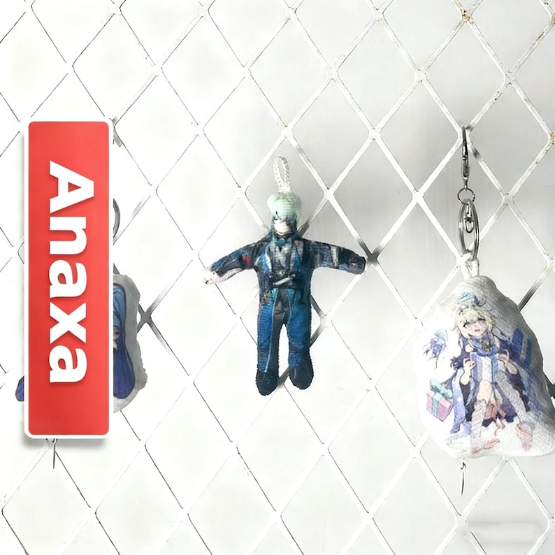 Op de afbeelding: Een rode rechthoekige tag met de witte tekst "Anaxa" wordt getoond naast twee sleutelhangers met personages. De sleutelhangers tonen personages in anime-stijl in blauwe en witte outfits. De achtergrond is een wit ruitvormig rooster.