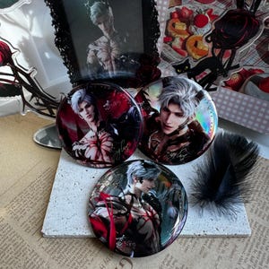 May include: Three round pinback buttons featuring a male anime character with white hair and red accents. The buttons have a glossy finish and are arranged on a textured surface with a feather and a framed picture in the background.