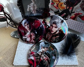 Puede incluir: Tres botones redondos con un personaje de anime masculino con cabello blanco y detalles rojos. Los botones tienen un acabado brillante y están dispuestos sobre una superficie texturizada, con una pluma y una foto enmarcada en el fondo.