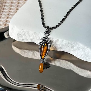 May include: A black metal pendant necklace with an amber-colored, teardrop-shaped stone. The pendant has an ornate, filigree design and hangs from a beaded chain. The necklace is displayed on a white, textured surface.