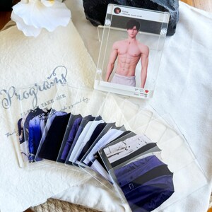 May include: A collection of clear acrylic cards featuring images of clothing and a shirtless man. The cards are arranged on a white cloth with the word "Programs" printed on it. The cards showcase various outfits.