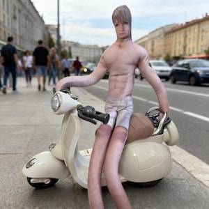 May include: A plush doll with a human face and torso, wearing shorts, sits on a cream-colored scooter. The doll has long arms and legs, and is posed on a city street with pedestrians and cars in the background.
