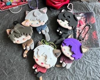 May include: A collection of cat-eared character keychains in various colors, including gray, black, white, brown, and purple. Each keychain features a cartoon-style design with a small loop for attachment. A red rose and a clear card are also visible.