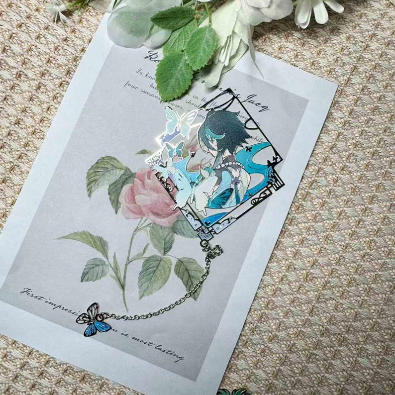 May include: A decorative bookmark featuring a character illustration with blue and teal accents, adorned with butterfly motifs. The bookmark is connected to a chain with a blue butterfly charm. The background includes a floral print and text.