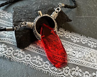 May include: A red crystal pendant necklace with a silver-colored metal setting and a black braided cord. The crystal has a faceted shape and a lace-like pattern within. The necklace is displayed on a dark gray stone.