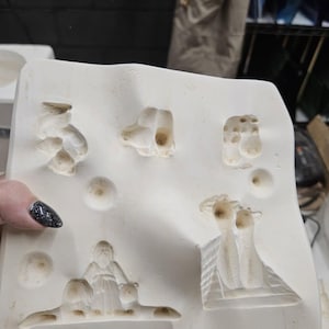 May include: A white plaster mold with various relief designs. The mold features several small, abstract shapes, along with a depiction of a figure with two animals. The mold is likely used for creating decorative objects.