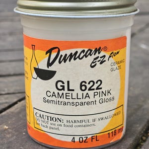 May include: A white ceramic glaze jar with a gold lid and an orange label. The label reads "Duncan E-Z Flow Ceramic Glaze GL 622 Camellia Pink Semitransparent Gloss." The jar contains 4 fl oz (118 ml).