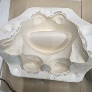 May include: White ceramic mold of a frog with a wide smile and large eyes. The mold is designed for making a 3D frog sculpture.
