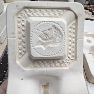May include: A white ceramic mold with a detailed relief design. The central square features a profile image within a dotted circle, surrounded by ornate patterns. The mold is likely for creating decorative items.