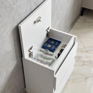May include: A white bedside cabinet with an open top compartment revealing a passport, watch, cash, and a black belt. The cabinet has two drawers and is set against a textured gray wall.
