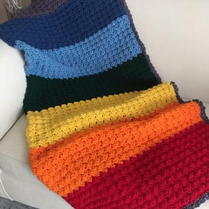 May include: A handmade, colorful crochet blanket draped over a white sofa. The blanket features a rainbow of colors, including red, orange, yellow, green, blue, and purple. The textured stitches and vibrant hues create a cozy and inviting aesthetic.