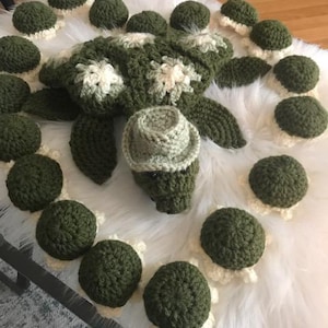 May include: A crocheted green turtle with a white hat and a white and green shell. The turtle is surrounded by smaller crocheted turtles in various shades of green.