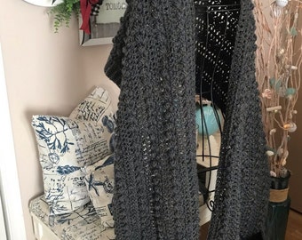 May include: A gray crocheted shawl with two pockets and fringe. The shawl is hanging on a black metal hanger. The text "Ellie Angel Hug Shawls" is visible on a sign in the background.