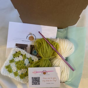 May include: A craft kit in an open cardboard box. The kit includes a white and green crocheted square, green and white yarn, a pink crochet hook, and a card that says "Scan me to open my shop on Etsy!" and "Thank You!"