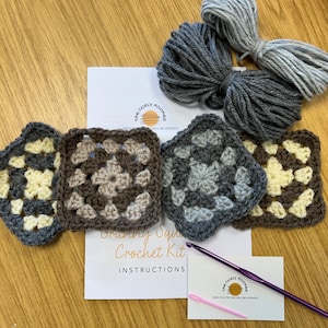 May include: A crochet kit with instructions for making granny squares. The kit includes four different coloured squares, two balls of yarn, and a crochet hook. The squares are made with brown, grey, and cream yarn.