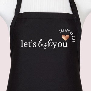 Puede incluir: Un delantal negro con el texto en blanco let's lash you y un pequeño icono de corazón de color cobre sobre la palabra you, con el texto adicional LASHED BY ELLE arqueado sobre el corazón. El delantal se muestra sobre un maniquí blanco.