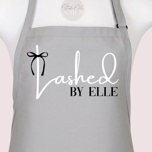 Puede incluir: Un delantal gris con un estampado gráfico en blanco y negro en la parte delantera. El texto dice "Lashed BY ELLE" con un pequeño icono de lazo negro sobre la letra L. El delantal tiene una correa para el cuello ajustable con hebilla negra.