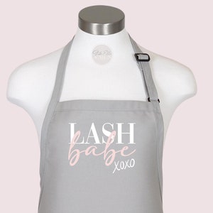 Puede incluir: Delantal gris con el texto "LASH babe xoxo" en letras blancas y rosas.