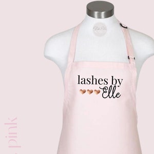 Puede incluir: Un delantal rosa con el texto "lashes by Elle" en letras negras y cuatro corazones dorados.
