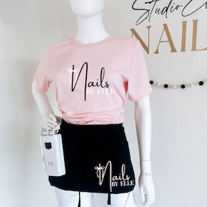May include: A pink t-shirt with the text "Nails by Elle" in black and white lettering. A black apron with the same text is worn over the shirt. The apron has a pocket on the side with a white electric nail drill.