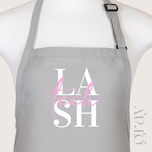 Puede incluir: Delantal gris con texto blanco que dice "LASH TECH" en una fuente estilizada. La palabra "TECH" está en rosa.