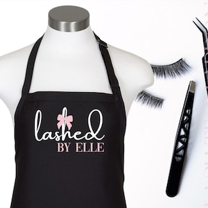 Puede incluir: Un delantal negro con el texto blanco "lashed", un pequeño lazo rosa sobre la letra "h" y el texto "BY ELLE" en rosa debajo. Junto al delantal hay dos pinzas de metal negro y un par de pestañas postizas de color negro.