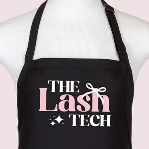 Puede incluir: Delantal negro con texto blanco que dice "The Lash Tech" con un lazo rosa y un diseño de estrella rosa.