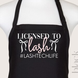 Puede incluir: Delantal negro con texto blanco que dice "Licensed to Lash #LASHTECHLIFE". Dos lazos rosas están a cada lado de la palabra "Lash".