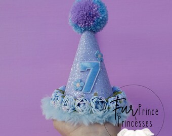 May include: A light blue glitter party hat with a blue pom-pom on top. The hat is decorated with blue flowers and a blue felt number 7. The hat is held in a hand.