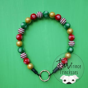 Op de afbeelding: Een feestelijke hondenhalsband met rode, groene en gouden kralen. De kralen zijn versierd met sneeuwvlok- en streepmotieven. De halsband heeft een goudkleurige metalen ring en zwarte connectoren. De achtergrond is effen groen.