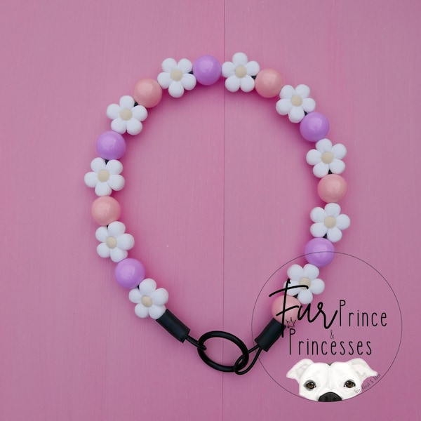May include: A pink and purple daisy flower beaded dog collar with a black metal clasp. The collar is decorated with white daisy flowers and pink and purple beads.