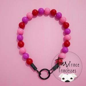 May include: A dog collar made of red, pink, and purple beads. The collar has a black metal ring and connectors. The background is a solid pink colour. The collar is a pet accessory.