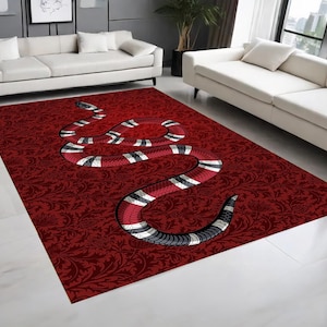 Pode incluir: Um tapete retangular com um fundo vermelho profundo e um padrão floral damasco tonal. Um gráfico grande de uma serpente sinuosa está centrado no tapete, exibindo faixas alternadas de vermelho, preto e branco ao longo de todo o seu corpo.