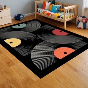 May include: Black rectangular area rug featuring a graphic design of vinyl records. The records have gray concentric circles with yellow, teal, red, and orange centers. The rug is placed on a wooden floor.