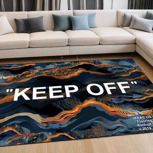 May include: A rectangular rug with a dark blue and orange abstract design, featuring the text "KEEP OFF" in large white letters. The rug is placed in a living room setting, with a cream-colored sofa and cushions in the background.
