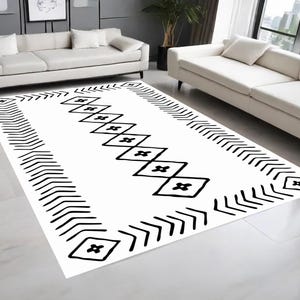May include: A white rectangular rug with a black geometric design. The pattern includes a central line of diamond shapes and an arrow-like border. The rug is in a living room with white sofas and a window.