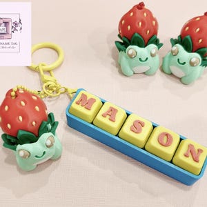 May include: A novelty keychain featuring a red strawberry top and a green frog-like body. The keychain has a yellow ring and a blue block with the name "MASON" in yellow and red letters. Two similar figurines are also present.