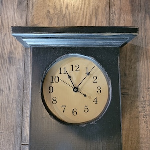 May include: A black wooden wall clock with a light-colored clock face. The clock has black hands and a black frame. The clock face displays the numbers 1 through 12. The clock is rectangular with a shelf at the top.