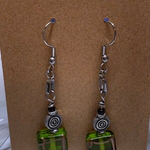 May include: Dangle earrings with silver hooks. Each earring has silver beads and rectangular accents, leading to a decorative silver bead and a square, transparent, lime green glass bead with pink accents.