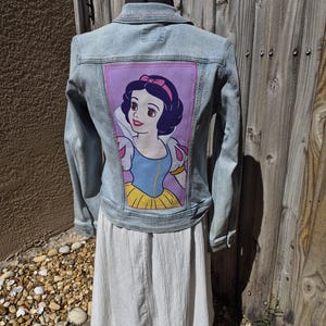 May include: Light blue denim jacket with a Snow White graphic on the back. The Snow White image is framed in a purple rectangle. The jacket is displayed on a mannequin with a white skirt.