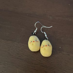 May include: A pair of novelty earrings featuring small, yellow potato-shaped charms with smiling faces. The earrings have silver-colored hooks and black accents, creating a whimsical and playful accessory.